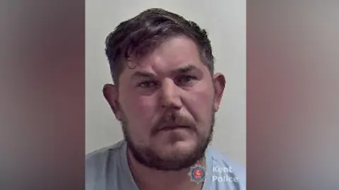 A mugshot of a man wearing a light blue top. He is looking directly at the camera.
