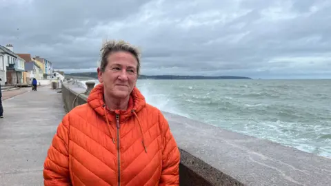 A woman with short blonde hair. She is wearing an orange coat. She is stood next to a sea wall. The sea is rough. The sky is grey with clouds. 