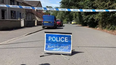 Blue police tape and a blue police sign saying 'road closed' blocks off the road past terrace houses. There is a police car parked in the distance, a blue van and red car parked up on the footpath.