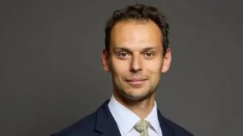 UK Parliament A man with short, dark, curly hair looks at the camera, wearing a navy jacket, white shirt and a pale green patterned tie.