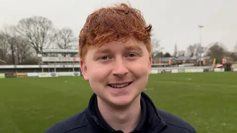 Robin Dyson, a young man with slightly curly ginger hair, is pictured smiling at the camera. He is stood in front of an empty football pitch.