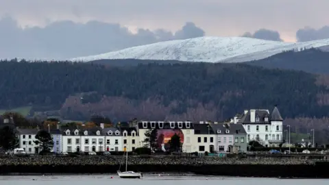 Ann Bruen A white beaked hill sits above a green tree line that sits above the town of warrenpoint. A row of white houses sit infront of the coast, a boat is in the harbour