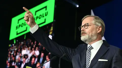 Michal Cizek/AFP man wearing a dark jacket and striped tie indicating his right hand to the public with the slogan of the green Spolu alliance behind his hand.