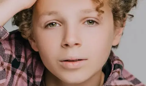 A professional-style headshot of a young boy looking into the camera while pushing a mop of curly hair away from his forehead.