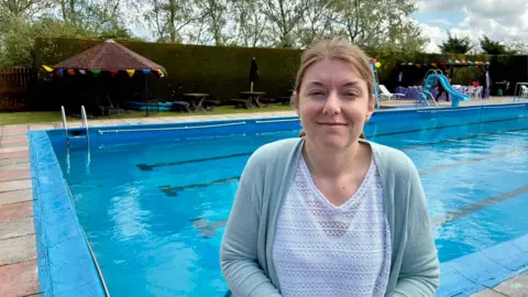 Debbie wears a white top and light blue cardigan and smiles for the camera in front of the pool.  A sun umbrella in the background has some coloured bunting on it.