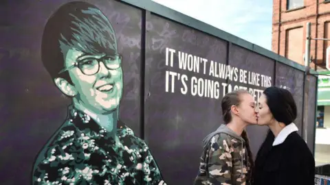Getty Images Robyn Eswards-Peoples (left) is wearing a camouflage jacket and grey hoodie. Sharni Edwards-Peoples (right) is wearing a black coat. They are kissing in front of the Lyra McKee mural in Belfast. Lyra is seen smiling and is wearing a colourful shirt. She is beside a quote reading: It won't always be like this. It's going to get better'
