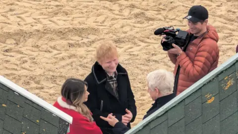 Amanda Jane Ed Sheeran - a man with red hair in a black jacket - stands on the beach next to a woman in a red jacket and a man with a camera and a cap. They talk to an older man with gray hair. 