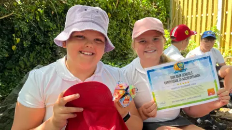 Pupils Charlotte, 10, and Hattie, 9, smiling wearing school uniform and sun hats. They are holding a sun hat and certificate. 