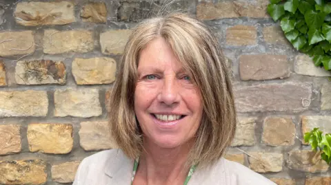 County Councillor Gina Dowding standing in front of a sandstone brick wall. In the far right corner there is a green plant. She is wearing a green dress with a beige blazer and is smiling.