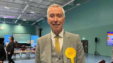A slim man in his sixties wearing a grey suit and waistcoat with a yellow tie smiling at the camera. He is in a sports hall, with tables and people behind him.