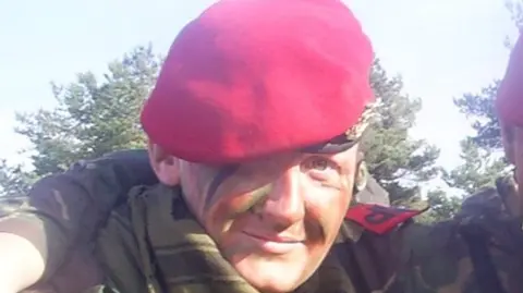 Zoë Robson A woman in a red beret, with smears of camouflage paint on her face, and wearing a camouflage top, looks at the camera