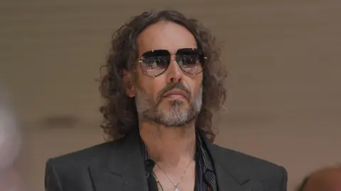 PA Media A headshot of Russell Brand wearing a grey blazer and sunglasses.