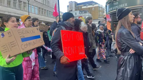 Protesters march ⁢with signs demanding rent cuts⁣ and highlighting "broken communities," and "cut our rent"