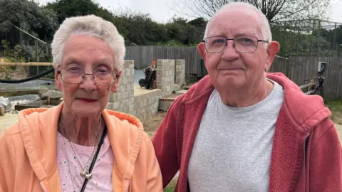 BBC News A couple stand next to each other at a seal sanctuary. On the left, the woman is wearing a peach jacket and pink top. The man wears a red jacket and grey top. They both wear glasses. 