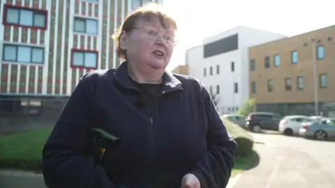 Alison Aitken, wearing a blue fleece and carrying a walking stick under her arm, speaks to a BBC reporter with the block of flats in the background