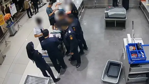 Group of security officers with faces blurred holding plastic bags, man dressed as pilot stands opposite them