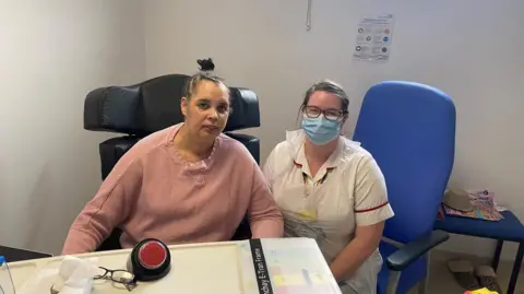 Two women sit down in hospital chairs and look at the camera. The woman on the left, Kerry Williams, wears a pink jumper and has her hair in a bun. The woman on the right wears a white nurse's uniform and a blue medical face mask.