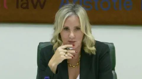PA Caroline Dinenage - blonde hair , red nails & holding a pen, dark jacket, gold chian sitting in a chair