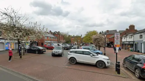 A street car park with a number of cars parked on a paved area surrounded by trees and buildings