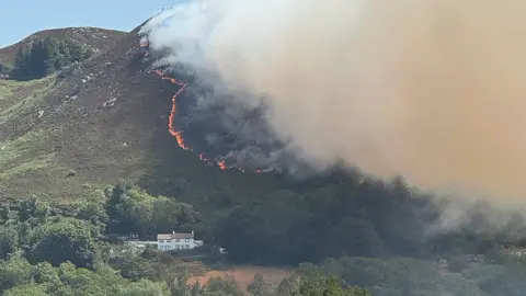 Rita Raweily A wildfire stretches across a wide area of mountainside in the direction of a home below