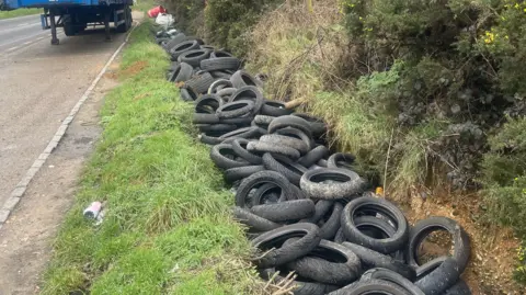 A ditch is filled to the brim with discarded tyres near a road.