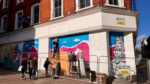 Street art being created on a building's exterior wall. The area has barriers to allow the artists to work. Shoppers walk past. It is sunny. 