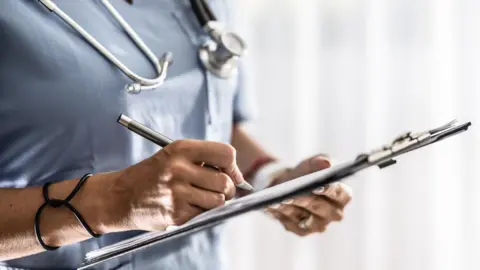 Getty Images A health professional wearing a blue uniform is using a pen to write on a sheet on a clipboard. They have a silver and black stethoscope around their neck and a black bracelet on their wrist. They are wearing a ring on their left hand. The background is blurred.