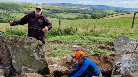 LDNPA Two people work to measure and place the slate shards in the ground, which is dug up. The terrain is in a farming area in Jackson Ground, the Lake District.