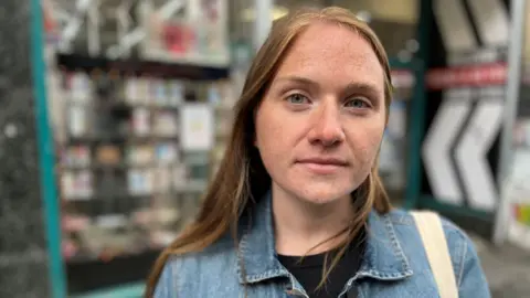 Laura Young, in a denim jacket over a black top, standing outside a shop with vapes displayed in the window.