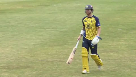 Ben Stokes on the pitch wearing a blue helmet and yellow and blue cricket kit and holding a bat in his right hand. He is walking to the left of the camera.