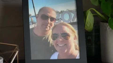 A photo frame containing a photo of John and Jo in sunglasses smiling to the camera in front of a sunny scene.