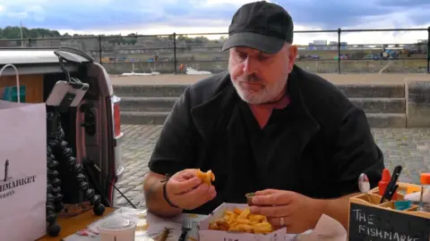 A man in black clothes and a black cap sits at a table outside eating fish and chips. A phone attacked to a tripod is on the table beside him. 