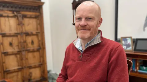 BBC Deputy Gavin St Pier sitting down wearing a red jumper