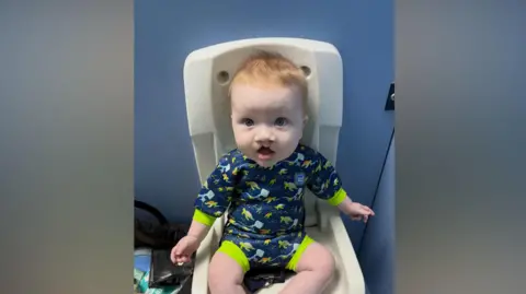 A baby, Eli. Pictured on a highchair, he is wearing a navy swimming costume that has turtles on it.