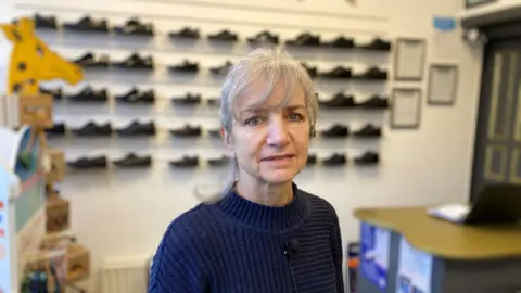 A woman looks into the camera. She is standing in the centre of a shop, with rows of children's shoes on a wall behind her. She has silver hair and wears a blue jumper. 