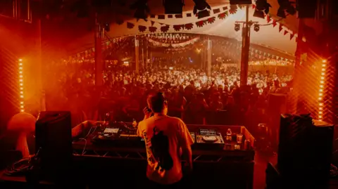 Shindig Festival A DJ performs in front of a large crowd of people inside a marquee which is lit up red by lights, at the Shindig Festival