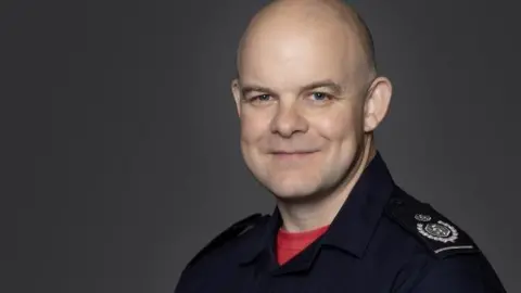 Formal work photo of Jonathan Smith, a bald man wearing a red T-shirt under a navy fire brigade boiler suit. There is a plain background and he is smiling.
