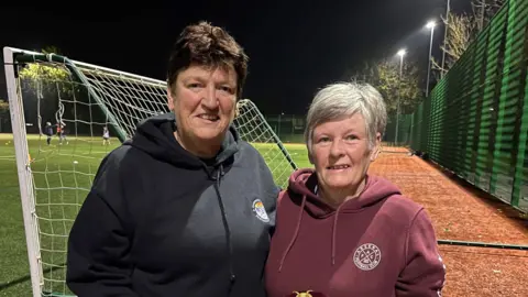 Mandy and Colleen wearing hoodies and standing in front of a football pitch smiling