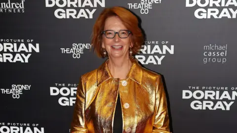 Julia Gillard attending an award ceremony. She has black glasses on and is wearing a gold jacket over a black top. Behind her is a black backdrop with the words The Picture of Dorian Gray in white, along with Sydney Theatre Co and Michael Cassel group. 