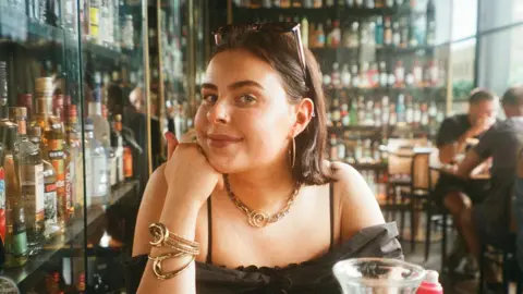 Abi A young woman in a bar with lots of bottles of drink in cabinets behind her. You can see the top half of her body. She is wearing a black top and resting her chin on her hand. She has sunglasses pinning back her hair and has golden earings, necklace and bracelet. 