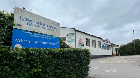 The sign for Lydney Hospital is visible with a no smoking sign below it. In the background is one of the buildings in the hopital, covered with hoardings of the site's marketer