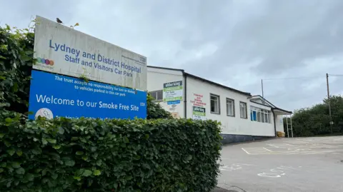 BBC The sign for Lydney Hospital is visible with a no smoking sign below it. In the background is one of the buildings in the hopital, covered with hoardings of the site's marketer