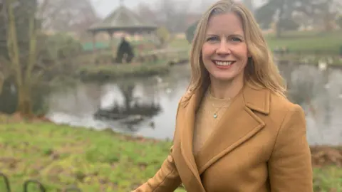 A woman wearing a tan coat and beige jumper stands in a park, smiling at the camera.