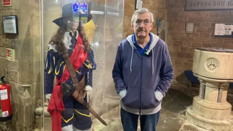 A man with short grey hair, a blue hooded jumper and jeans, stands with his hands in his pockets. Next to him is a model of a Civil War-era soldier, encased in a glass box.