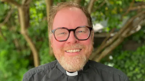 The Reverend Matthew Simpkins has short fair hair and is smiling in front of a green bush. He is wearing his vicar uniform, which consists of a black shirt with a white collar.