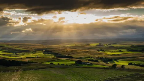 Luke_Northumberland A valley of green fields interspersed with fences, trees and hedges. The sky is covered in dark clouds, casting the fields in shadow. Partway up the sky is a break in the clouds with the bridge sun coming through. Beams of light spread out from the sun, lighting parts of the shadowed fields.
