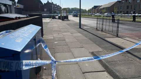 BBC Police tape surrounds an paved area close to a pub and a shop. Two police officers stand close to the cordon.