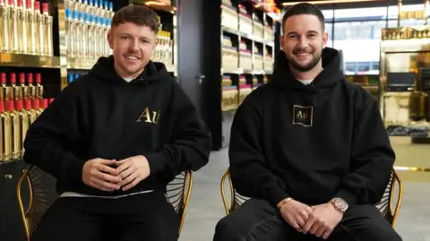 Two young men are smiling as they sit in golden framed chairs next to a row of gold-coloured Au Vodka bottles. They are both wearing black hoodies with the gold AU Vodka logo on them and black trousers. 