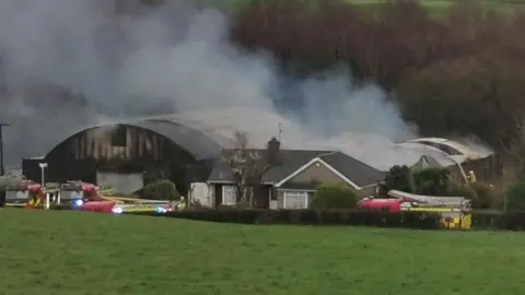 Over the brow of a green field, red and yellow fire engines can be seen in front of a farm house. Behind the farmhouse, smoke bellows from a collection of sheds. 
