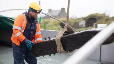 Orkney Islands Council A bearded man in an orange hi vis jacket places a large timber beam into a water tank he is standing in